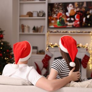 a man and woman in Santa hats watching a Christmas movie by a tree and fireplace